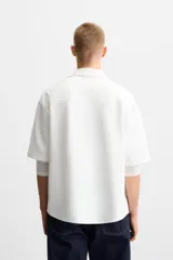 Sobrecamisa color beige de tejido elástico, con cuello solapa, manga corta y bolsillos de parche en el pecho. Cierre frontal con botones a presión.