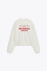 Sudadera color crudo de cuello redondo y manga larga, con estampado frontal en color rojo con texto "Sunset view Await", "San Francisco" y "Golden Gate".