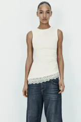 Camiseta ajustada (slim fit) de cuello redondo y manga sisa, color beige claro, con frunces laterales y bajo asimétrico con terminación de encaje a tono.