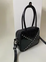 Cartera rígida negra de cuero con tachas plateadas y tres combinaciones de cuero diferentes que aportan textura. Tiene herrajes y cierre metálico plateado marca YKK. Cuenta con un asa fina para llevar al hombro o en la mano y una correa larga extra regulable.