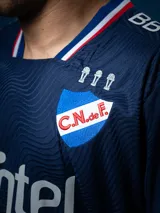 Camiseta de fútbol Umbro del Club Nacional de Football, modelo Away 2025. Diseño azul con detalles en blanco y rojo en cuello y mangas. Incluye logo de Umbro, escudo del club y sponsor Antel en el frente.