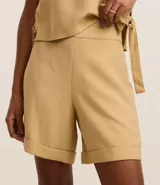 Bermuda femenina de corte recto y cintura alta, color beige, confeccionada en viscosa twill. Presenta abertura lateral por cremallera y ruedo plegado hacia afuera.