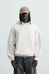 Sudadera con capucha color crudo, de corte relajado, confeccionada en tejido de algodón con interior rizado. Cuenta con cuello con capucha, manga larga, bolsillo frontal tipo canguro y acabados en rib.