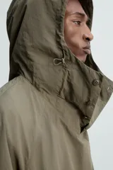 Parka color verde militar de corte amplio, confeccionada en tejido técnico ligero. Cuenta con cuello alto con capucha ajustable, cierre frontal con botones, mangas largas con puños ajustables, bolsillos de plastrón con solapa en la parte delantera, cintura ajustable con cordón y bajo con aberturas laterales ajustables con cordón.
