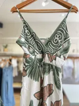 Vestido largo con breteles finos, escote en V y estampado floral en tonos verdes y marrones sobre fondo blanco. Tiene lazo ajustable en la cintura.
