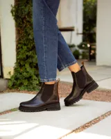 Bota tipo Chelsea de cuero negro con paneles elásticos laterales. Presenta una suela track de goma y un tirador de cuero marrón en la parte posterior.