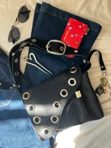 Cartera bandolera de cuero negro con ojalillos metálicos plateados en el frente y correa regulable con hebilla. Tiene un bolsillo exterior y gancho interior para llaves.