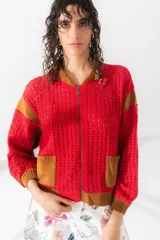 Chaqueta estilo bomber tejida de punto calado color rojo con detalles en marrón en cuello, puños, ruedo, mangas y bolsillos. Tiene cierre frontal y apliques de flores en el pecho.