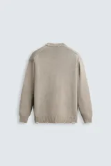 Jersey de punto color beige, con cuello redondo, manga larga y corte holgado. Presenta un lavado con efecto desgastado y rotos en toda la prenda.
