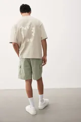 Short cargo color verde claro en popelina de algodón, con cintura elástica ajustable con cordón, bolsillos laterales, bolsillos cargo con solapa y botón a presión en piernas y traseros, y aberturas laterales en el bajo.