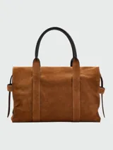 Bolso tipo tote de gamuza color marrón, con forma abombada y correas ajustables con hebillas doradas. Presenta asas de hombro en cuero negro y detalles de costura visibles.