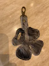 Llavero con forma de trébol de cuatro hojas, confeccionado en cuero con pelo en tonos gris y negro. Incluye un gancho metálico tipo mosquetón en tono bronce y una pequeña placa metálica grabada con la marca "SHAW".