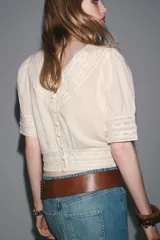 Blusa color crudo de cuello redondo y manga corta con puños. Presenta bordados a tono y cierre trasero con botones forrados.