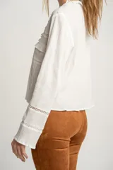 Blusa blanca de algodón con escote en V, cierre con lazos, mangas largas con puños elásticos y detalles de encaje y volados.