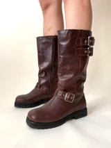 Bota de caña media de cuero vacuno color marrón oscuro, con punta cuadrada, cierre lateral y suela track negra. Presenta detalles de hebillas metálicas plateadas en la caña y en la parte inferior del empeine.