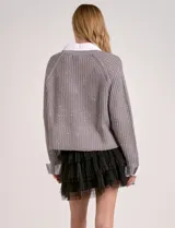 Buzo de tejido de punto gris, con cuello en V y detalles de lentejuelas. Se lleva superpuesto a una camisa blanca con cuello y puños visibles.