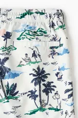 Short de baño blanco con estampado de palmeras, personas y paisajes en tonos azul, verde y rosa. Tiene cintura elástica con cordón ajustable, bolsillos laterales, bolsillo trasero con velcro y suspensor interior de malla.