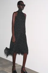 Vestido midi negro con estampado de lunares blancos, cuello halter con pañuelo integrado, sin mangas y bajo asimétrico. Cierre con abertura y botón en la nuca.