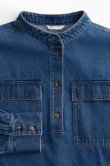Camisa de denim azul de corte holgado, con cuello mao, tapeta de botones, hombros caídos, mangas largas con puños abotonados y dos bolsillos superiores con solapa.