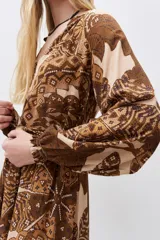 Vestido largo de viscosa estampada en tonos marrón y beige, con escote en V, mangas largas con puños elásticos y volado en el ruedo. Incluye lazo del mismo género para ajustar en la cintura.
