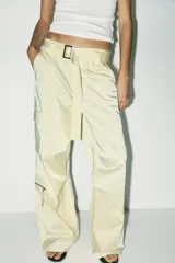 Pantalón cargo color blanco hueso, de tiro medio, corte ancho y tela satinada. Incluye cinturón con hebilla metálica, bolsillos delanteros, laterales con cierre y traseros de vivo. Cierre frontal con cremallera y ganchos metálicos.