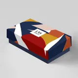 Billetera corta de símil cuero color beige claro, con cierre de broche a presión en la solapa principal. Presenta múltiples compartimentos internos para tarjetas y billetes, y un bolsillo exterior con cierre en la parte posterior. Lleva una placa metálica con el logo de la marca.