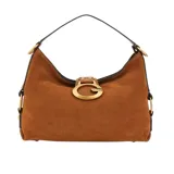 Bolso de hombro tipo hobo de gamuza color marrón coñac, con correa delgada negra y herrajes dorados. Presenta un gran dije metálico con el logo 'G' de Guess en el centro frontal.