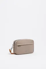 Bolso bandolera mini de nylon color beige, con estructura blanda, cierre de cremallera y asa regulable de tejido. Presenta un bolsillo exterior frontal con ojal metálico y el logo metálico de Bimba y Lola en la parte inferior.