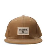 Gorro tipo snapback color marrón claro con visera plana y costuras blancas. Lleva un parche rectangular frontal de tela cruda con el logo y texto "KRISTOBELGA ESTD. SINCE 1981 - K - SURF & SKATE GOODS".