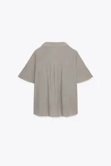 Camisa color gris claro con cuello solapa y escote pico, de manga corta. Presenta un nudo en la parte delantera y cierre frontal con botones.