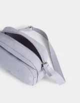 Bandolera gris claro de tela deportiva con bolsillo frontal, correa larga regulable, acceso con cierre superior y tag de goma.