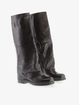 Bota alta de cuero bovino color negro con diseño de caña ancha y efecto doblado. Presenta detalles de tachas metálicas a lo largo de la costura posterior y una placa metálica decorativa en la parte superior. Cuenta con tacón bajo y punta redondeada.