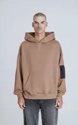 Buzo hoodie marrón de corte oversize, confeccionado en algodón rústico. Tiene capucha forrada, bolsillo tipo bomber con cierre en la manga y bolsillo canguro.