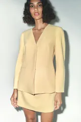 Blazer amarillo pálido de corte recto, con escote en pico y manga larga. Presenta bolsillos de ojal en el delantero y cierre frontal con botones a presión.