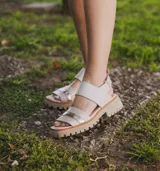 Sandalias de cuero color crudo con dos tiras horizontales en el empeine, una de cuero liso y otra con detalle dorado. Suela track color beige.