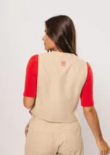 Chaleco beige con escote en V, sin mangas y cierre frontal con botones. Presenta pespuntes en color rojo y logo bordado en la espalda.