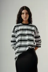 Camiseta de manga larga, corte holgado, con cuello redondo y estampado de rayas horizontales anchas en blanco y negro. Presenta un pequeño bordado rojo en el pecho.