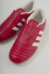 Championes Adidas rojos de cuero sintético, diseñados para taekwondo. Presentan el logo de Adidas en la lengüeta y las tres rayas blancas características en los laterales. El interior es acolchado y de color blanco.