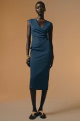 Vestido midi color beige, de corte recto, con escote en pico cruzado, sin mangas y detalle de hebilla metálica en la cintura.
