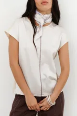 Blusa de manga corta color crema con acabado satinado y textura arrugada. Presenta un pespunte central vertical en color marrón oscuro y paneles laterales en tejido de punto color beige.