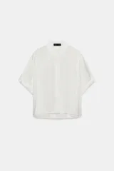 Camisa blanca de manga corta con cuello solapa y puños vueltos. Presenta un bajo asimétrico con aberturas laterales y cierre frontal oculto por solapa.