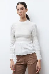 Blusa de color crudo con cuello alto fruncido y pechera con nido de abeja. Cuenta con mangas largas con detalles elásticos y puños con volados, y un bajo de corte peplum.