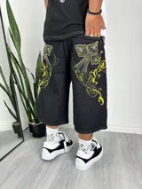 Bermuda de denim negro, corte muy ancho y largo (tipo culotte o capri), con bordados grandes en hilo amarillo y blanco en forma de cruces ornamentadas en los bolsillos delanteros.