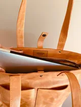 Cartera tote de cuero color tostado, con doble asa de mano y correa larga extraíble y ajustable. Tiene cierre superior y dos bolsillos externos. Interior forrado con compartimento acolchado para laptop de hasta 14 o 16 pulgadas.