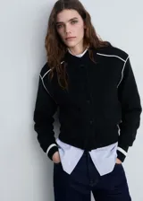 Chaqueta negra de punto con corte bomber, cierre frontal con botones a presión, bolsillos laterales y ribete blanco en cuello, puños, ruedo y hombros.