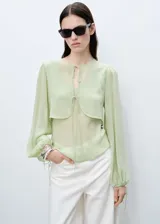 Blusa color verde claro de gasa semitransparente con cuello redondo y lazos en el frente. Tiene mangas largas abullonadas con puños elásticos y fruncidos. Incluye un chaleco corto incorporado. 