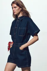 Mono corto de denim azul oscuro, con cuello solapa, manga corta, bolsillos delanteros y traseros, cintura elástica en la espalda y cierre frontal con cremallera y botones metálicos.