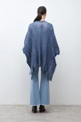 Ruana de tejido de punto en tonos beige y blanco, con patrón de espiga. Presenta cuello en V, mangas amplias, dos bolsillos frontales y flecos en el dobladillo.