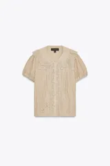 Camisa color crema con cuello bobo, manga corta abullonada con elástico y detalle de bordados combinados a tono y pliegues. Cierre frontal con botones forrados.