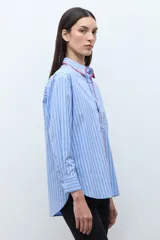 Camisa de manga larga, corte oversize, color celeste con rayas verticales blancas. Presenta un detalle de bordado en color rojo/fucsia a lo largo del cuello y la abertura frontal.